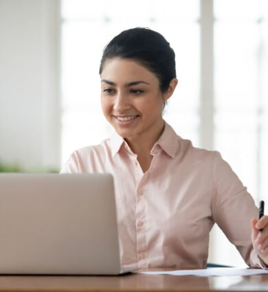 Employee Header Happy millennial Indian businesswoman sit at desk in office work online on laptop busy with paperwork. Smiling young mixed race female employee worker look at computer screen consult client distant.